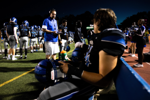 Eine Gruppe von Menschen auf einem Fußballfeld, einige tragen Helme und andere halten Flaschen, mit Bäumen und einem klaren blauen Himmel im Hintergrund.