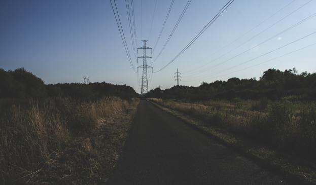 Eine Straße mit Stromleitungen, die entlang verlaufen, umgeben von saftigem Grün und Bäumen auf beiden Seiten, mit hohen Türmen und Drähten im Hintergrund vor einem klaren blauen Himmel.