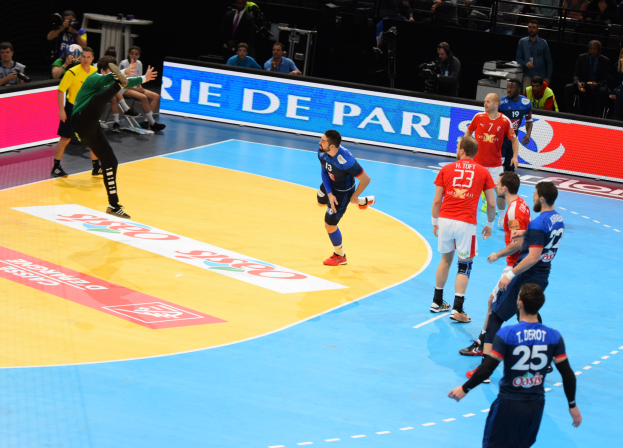 Eine Gruppe von Männern, die Handball auf einem Feld spielen, mit Zuschauern und Kameras im Hintergrund und Schildern mit Text, der auf die Futsal-Weltmeisterschaft 2019 hinweist.