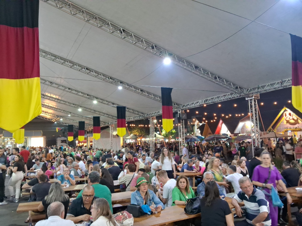 Eine große Gruppe von Menschen sitzt an Tischen unter einem Zelt beim Oktoberfest in München, Deutschland, mit anderen, die stehen und Gegenstände auf den Tischen, umgeben von Zelten, Lichtern und Fahnen.