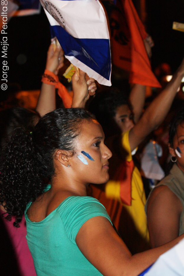 Eine Frau in einem grünen T-Shirt steht auf einer Demonstration und hält ein weißes Schild in der Hand, während viele andere Menschen hinter ihr stehen.