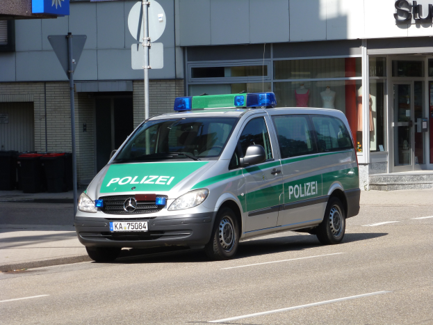 Ein Polizeiwagen parkt am Straßenrand vor einem Gebäude mit Glastüren und Schaufensterpuppen, mit Schildern und Mülltonnen im Hintergrund.