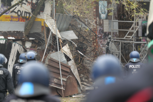 Eine Gruppe von Polizisten mit Helmen steht vor einem teilweise abgerissenen Gebäude mit Text an seiner Wand, umgeben von Bäumen und Masten.