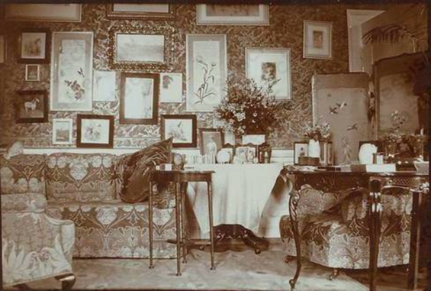 Ein altes Schwarz-Weiß-Foto eines Wohnzimmers mit einem Studio-Sofa, einem Stuhl und einem Tisch mit Blumenvasen, Fotorahmen und anderen Gegenständen, mit gerahmten Wänden, die eine gemütliche Atmosphäre schaffen.