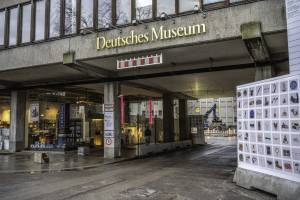 Außenansicht des Deutschen Museums in Berlin mit einem großen Glasgebäude, Säulen, einem Namensschild und einer Informationsanzeigetafel auf der rechten Seite, mit Bäumen und anderen Gebäuden im Hintergrund.