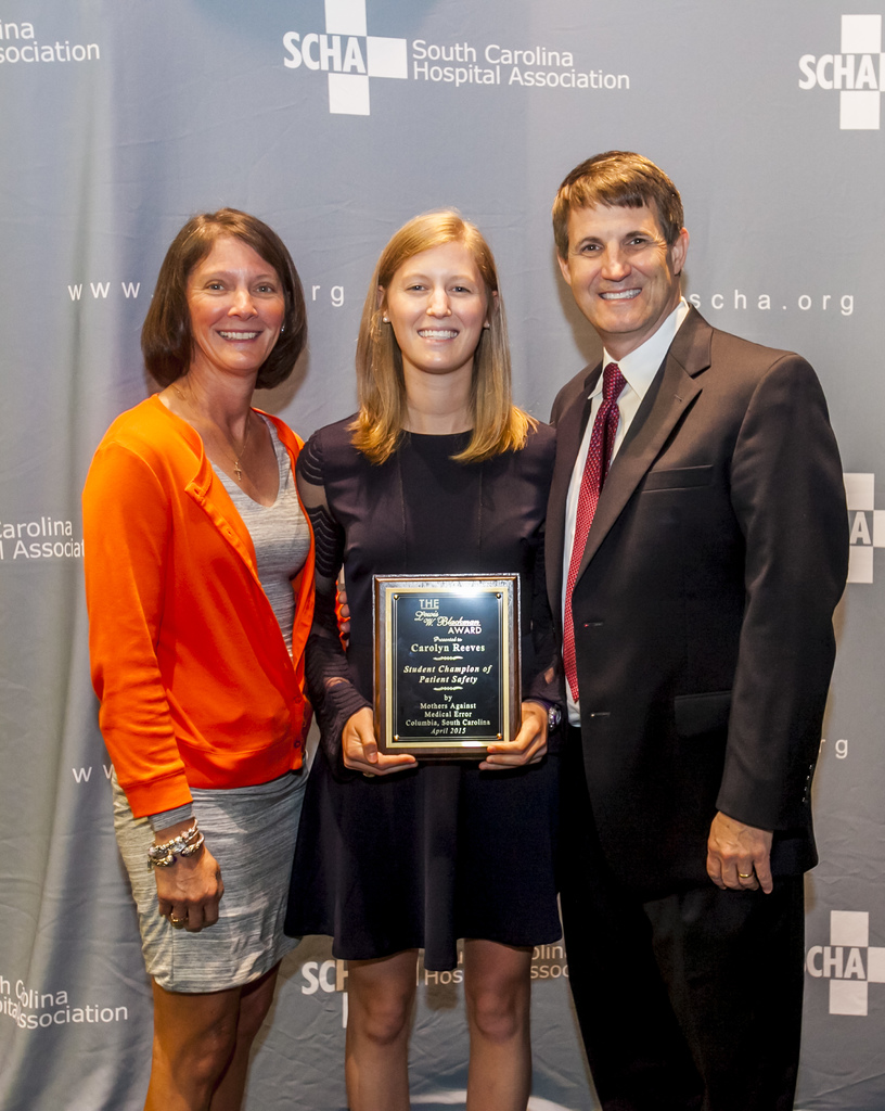 Drei Personen, zwei Frauen und ein Mann, stehen zusammen. Der Mann trägt einen schwarzen Anzug, eine Frau trägt ein orange-schwarzes Kleid und die andere Frau trägt ein schwarzes Kleid und hält eine Trophäe in der Hand.