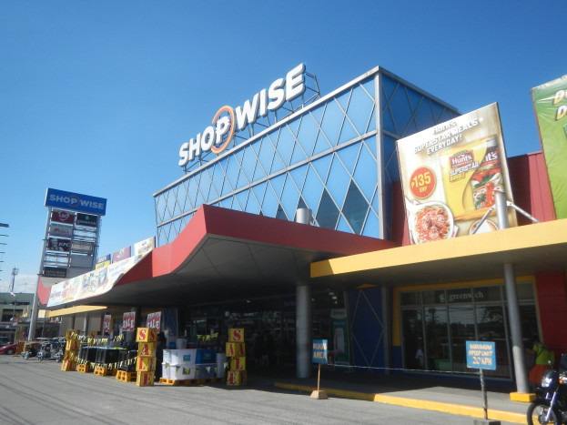 Ein Supermarkt namens Shopwise mit einem vor der Tür geparkten Motorrad, umgeben von Fahrzeugen, Pfählen, Plakaten und Werbetafeln auf der Straße, mit Gebäuden, Bäumen und einem klaren blauen Himmel im Hintergrund.