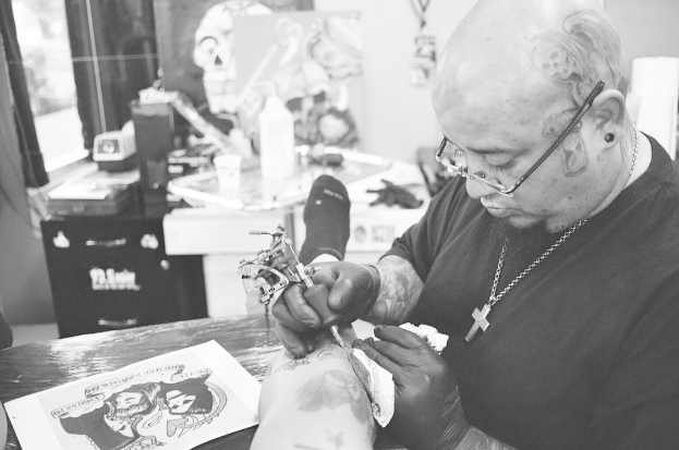 Ein Mann in einem schwarzen T-Shirt, mit Brille und einer Kette um den Hals tätowiert ein Bein mit einer Maschine, mit einem Papier auf der Oberfläche und einem unscharfen Hintergrund mit einer Flasche, einem Becher und anderen Gegenständen.