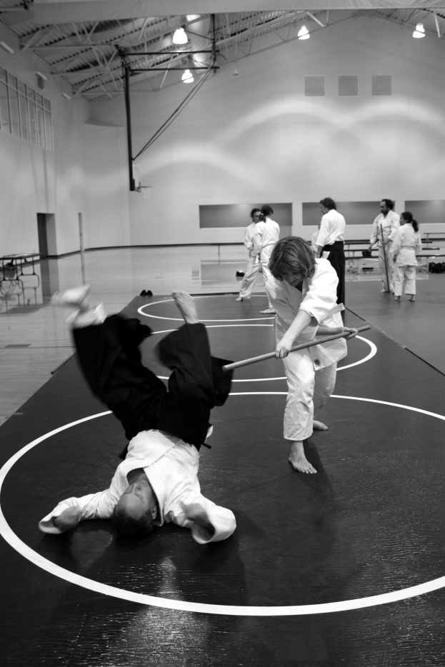 Zwei Personen üben Karate mit Stöcken in einer Turnhalle, während andere zuschauen, mit sichtbaren Lichtern an der Wand im Hintergrund.