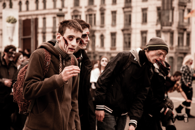 Eine Gruppe von Menschen in Zombie-Kostümen geht eine Straße entlang, mit einem Gebäude im Hintergrund, einer Person hält eine Kamera.