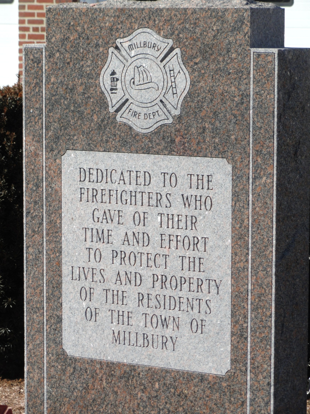 Gedenkstein mit einer Plakette für Feuerwehrleute in Millbury, vor einem Backsteingebäude mit umgebender Vegetation aufgestellt.