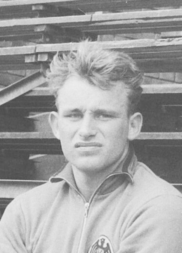 Schwarzes und weißes Foto eines Mannes in einer Jacke, der vor gestapelten Holzplanken steht, der legendären deutschen Füßballer.