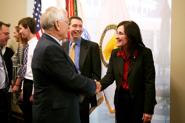 Ein Mann und eine Frau in Blazern und Hemden schütteln sich die Hand und lächeln sich an, während ein Mann in Blazer, Krawatte und Hemd neben ihnen steht; im Hintergrund unterhalten sich Menschen und lächeln in der Nähe von Fahnen an einer Wand.