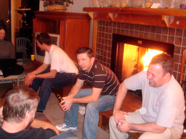 Vier Männer sitzen auf einer Bank, eine Frau sitzt links auf einem Stuhl, ein Feuer brennt im Kamin, ein Holzbrett hält Papier, einen Ball und Schaustücke, ein hölzerner Schreibtisch zeigt einen Blumentopf und einen Becher, mit einem Fernseher, Stuhl und Lampen im Hintergrund.