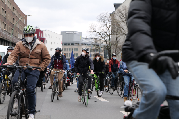 Eine Gruppe von Menschen in Helmen und Handschuhen fährt Fahrräder auf einer von Bäumen gesäumten Straße in Berlin, Deutschland, mit Gebäuden und einem geparkten Fahrzeug im Hintergrund.