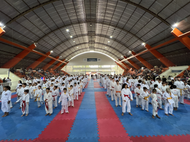 Eine große Gruppe von Menschen steht auf einer blauen und roten Matte in einer Turnhalle mit anderen, die auf Treppen sitzen und stehen, unter beleuchteten Decken und Tafeln mit Text im Hintergrund, die auf ein Taekwondo-Event hinweisen.