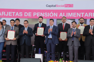 Eine Gruppe von Menschen auf einer Bühne, die klatschen und Zertifikate halten, mit Stühlen in einem Halbkreis und einem Banner mit der Aufschrift "Capital Social for Ti" im Hintergrund, sowie Blumensträuße und Redner am Boden.