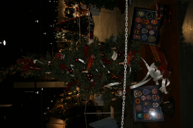 Ein Weihnachtsbaum mit Geschenken, Rahmen und Spielzeugen auf einer Plattform, mit Menschen, zusätzlichen Weihnachtsbäumen und Dekorationen im Hintergrund.