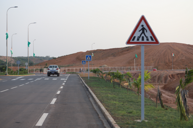 Ein Auto fährt auf einer Straße mit einem Fußgängerüberweg-Schild auf der Seite, flankiert von Laternenpfählen, Bäumen, Gras und einem Hügel mit klarem blauem Himmel im Hintergrund.