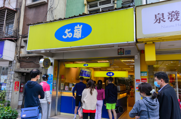 A group of people stand in front of a store with windows, signboards, plants, a fire extinguisher, and a roof with ceiling lights; some wear bags and masks, suggesting Covid-19 precautions.
