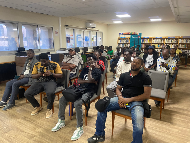 Eine Gruppe von Menschen sitzt in Stühlen in einem Raum mit Bücherregalen, einige halten Telefone und Taschen, andere sehen auf ihre Telefone, mit Monitoren und Gegenständen auf Tischen, Plakaten an den Wänden, Deckenleuchten, einer Klimaanlage und einer Tafel, scheinbar an einer Vorlesung über Bildung teilnehmend.