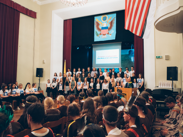 Eine Gruppe von Menschen steht an einem Rednerpult mit Mikrofon, flankiert von Fahnen und Lautsprechern, vor einem sitzenden Publikum, mit einem Bildschirm, der Text und ein Bild anzeigt, sowie Vorhängen und einem Kronleuchter im Hintergrund bei einem Kunst- und Kulturereignis.