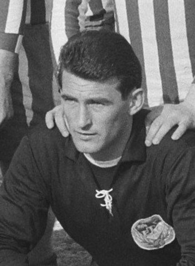 Schwarzes und weißes Foto eines Mannes, der auf einem grünen Fußballfeld sitzt und ein T-Shirt und eine Kette trägt, mit zwei Personen dahinter.