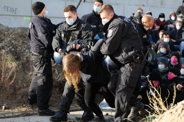 Eine Gruppe maskierter Personen in Kapuzenpullis, einige bewaffnet, umgibt eine Frau, die von der Polizei weggeführt wird, mit Pflanzen, Steinen und einer Wand mit Protesttext im Hintergrund.