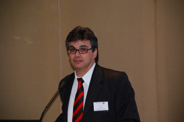 Eine Person in einem Anzug, die ein Abzeichen und eine Brille trägt, lächelt und vor einem Mikrofon auf einem Ständer steht, mit einer Wand im Hintergrund.