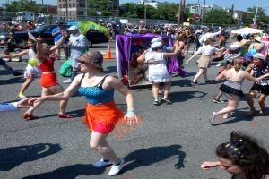Eine Gruppe von Menschen, die auf der Straße in einer Parade gehen, einige mit Mützen und Schirmen, andere tanzen, mit einem Banner, Verkehrskegeln, Fahrzeugen, Laternen, Schildern, Bäumen, Gebäuden und einem klaren blauen Himmel im Hintergrund.