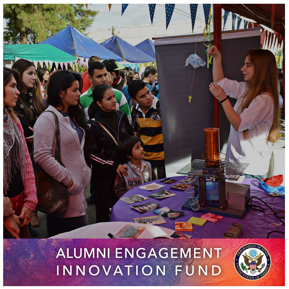 Gruppe von Menschen um einen Tisch versammelt mit einer "Alumni Engagement Innovation Fund"-Aufschrift, umgeben von Gegenständen, mit Zelten und Bäumen im Hintergrund unter einem klaren blauen Himmel, und Text und ein Logo unten.