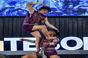 Zwei Performer in purpurfarbenen Kleidern führen einen akrobatischen Hebemanöver auf der Bühne vor einem Publikum aus, mit einem schwarzen Vorhang und einem Bildschirm im Hintergrund.