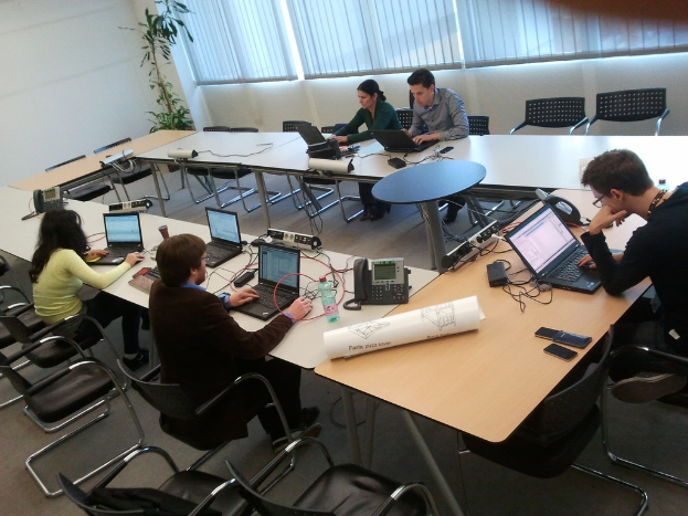 Fünf Personen sitzen an einem Tisch und arbeiten an Laptops, mit einem Telefon, einer Grafik, einer Fernbedienung und einem Handy auf dem Tisch; im Hintergrund ist ein Vorhang, eine kleine Pflanze und eine Wand zu sehen.