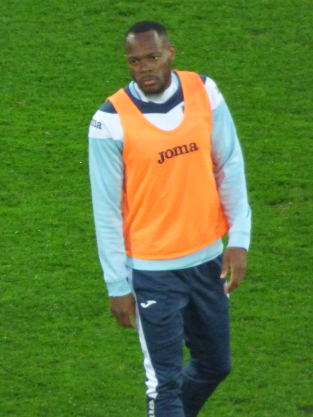 Ein Fussballspieler in einem orangen T-Shirt und blauen Hosen überquert entschlossen ein grünes Feld.