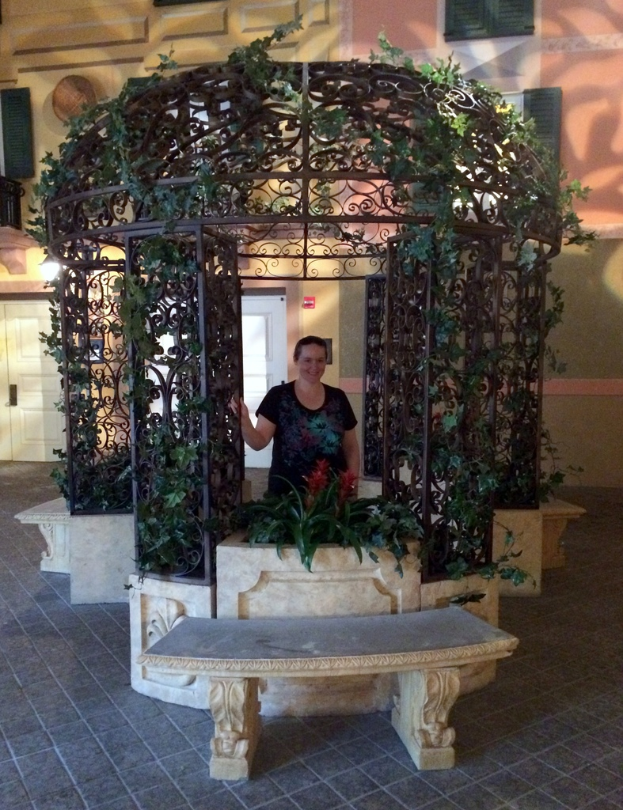 Eine Bank im Vordergrund mit einer verzierten Eisenpfeiler und Kletterpflanzen dahinter, mit einer Frau in einem schwarzen Kleid und einem Gebäude mit Wänden, Fenstern und Türen im Hintergrund.