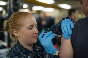 Eine Frau in militärischer Uniform erhält eine Impfung von einem Mann mit Handschuhen und Uhr, mit einem unscharfen Hintergrund.