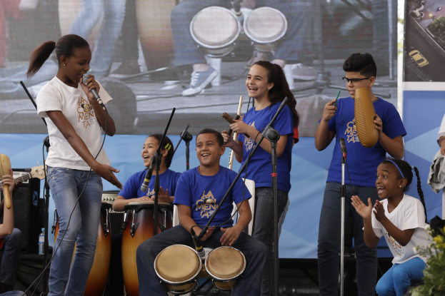 Kinder auf der Bühne mit Musikinstrumenten, eine Frau mit Mikrofon in der Mitte, Mikrofonständer, ein Banner im Hintergrund und Topfpflanzen rechts.
