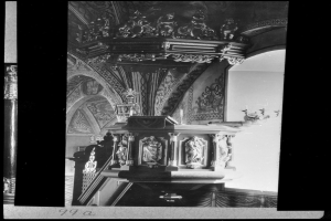 Ein Schwarz-Weiß-Foto eines Kirchenpults mit geschnitzten Säulen und einer strukturierten Wand im Hintergrund, das unten einige Text enthält.