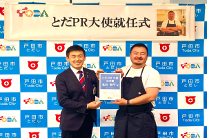 Zwei lächelnde Männer mit einem Zertifikat, im Hintergrund eine Tafel mit der Aufschrift "Toda City gewinnt PR Award" und einem Foto einer Person.