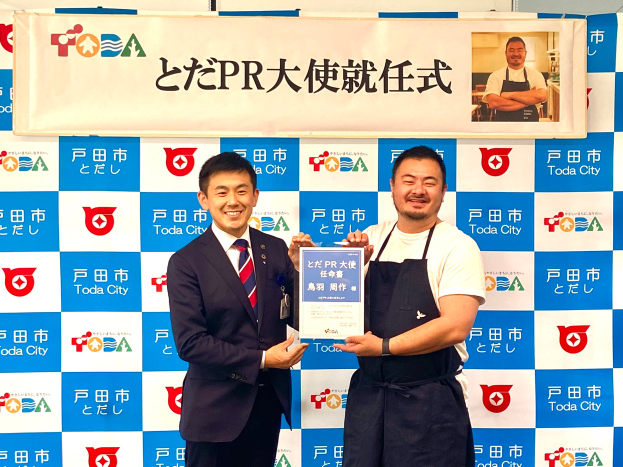 Zwei lächelnde Männer mit einem Zertifikat, im Hintergrund eine Tafel mit der Aufschrift "Toda City gewinnt PR Award" und einem Foto einer Person.