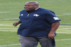 Ein Mann in einem blauen Hemd und grauen Hosen steht auf einem grünen Feld und lächelt mit einem selbstbewussten und entschlossenen Gesichtsausdruck, der den Eindruck eines Fußballtrainers erweckt.