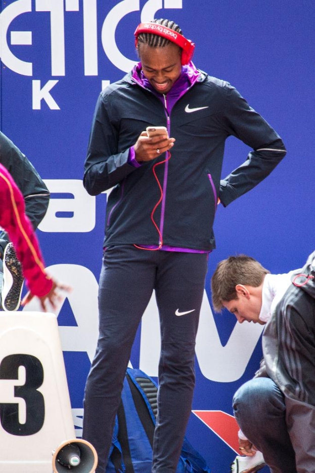 Eine Person, die mit einem Mobiltelefon in der Hand steht und lächelt, daneben eine weitere Person, die in der Hocke sitzt, ein Rucksack, ein Megafon unten, eine Wand im Hintergrund und eine weitere Person, die steht.