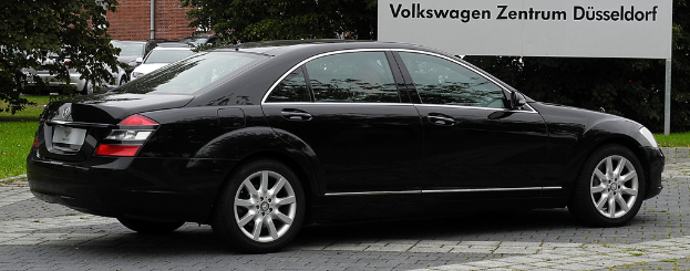 Mehrere Autos in der Nähe einer Wand geparkt, ein Rohr an der Wand befestigt, Bäume, Gras, Blätter auf dem Boden, ein schwarzes Auto in der Nähe einer weißen Tafel mit Text an Pfählen.