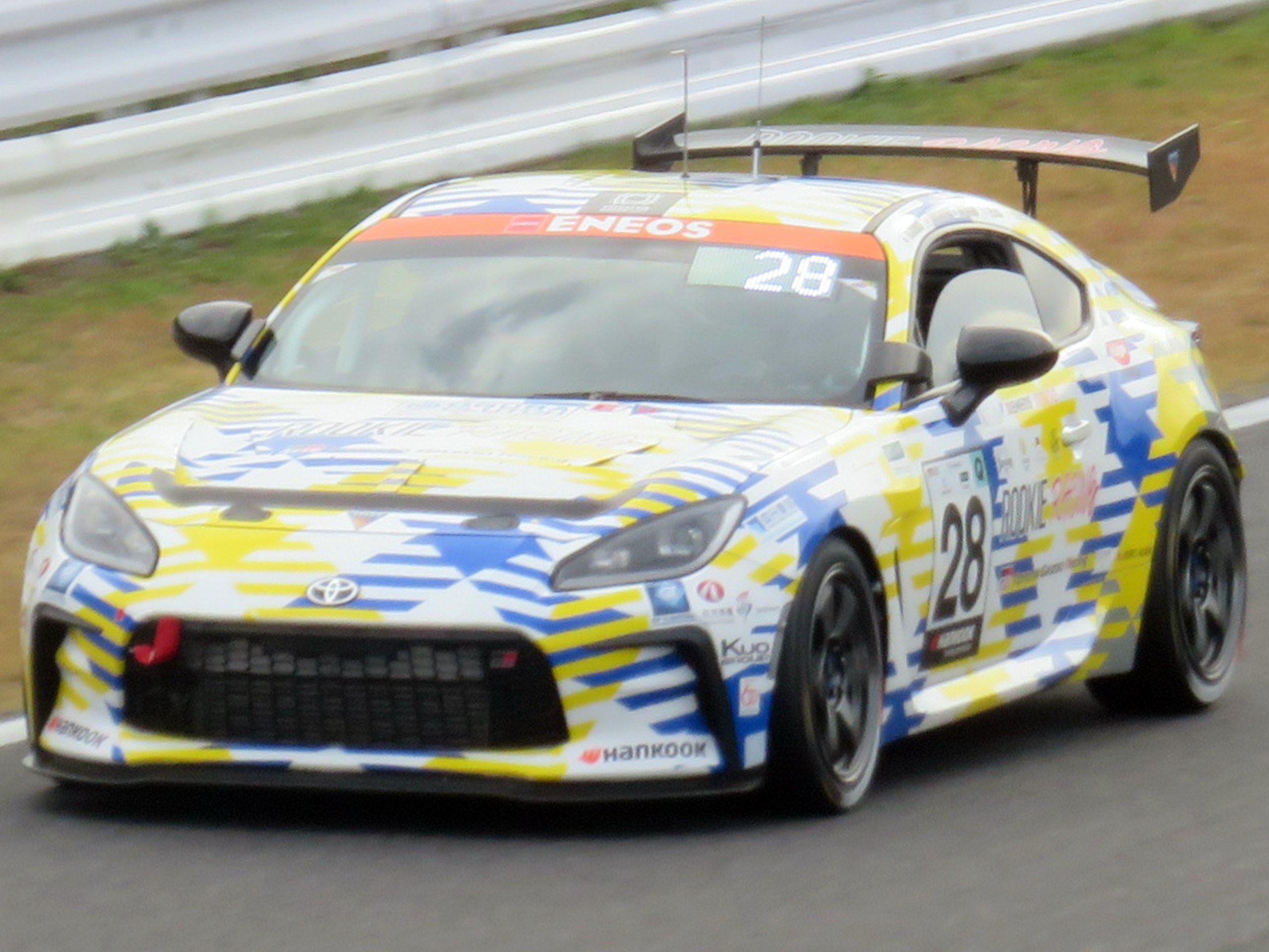 Ein weiß-blaues Toyota Supra GT3-Rennfahrzeug auf einer Rennstrecke mit Sponsoren-Logos, einer Begrenzung und Gras im Hintergrund.