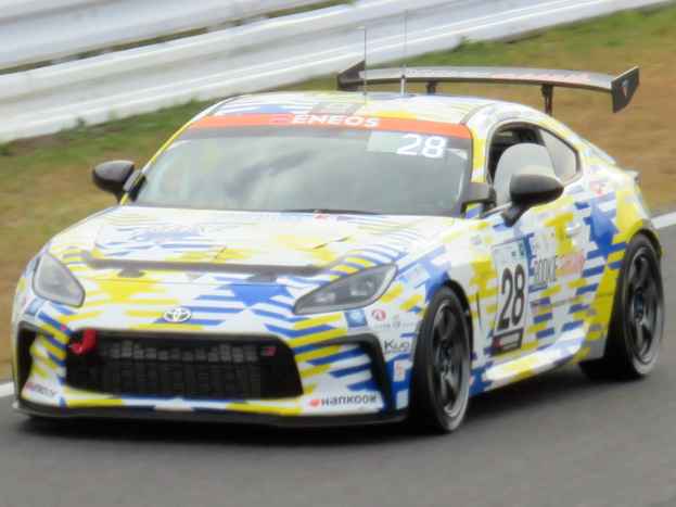 Ein weiß-blaues Toyota Supra GT3-Rennfahrzeug auf einer Rennstrecke mit Sponsoren-Logos, einer Begrenzung und Gras im Hintergrund.