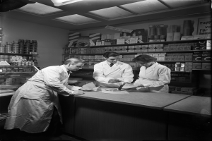 Drei Männer in weißen Kitteln stehen an einem Tresen in einer Apotheke mit Papieren und Gegenständen darauf, mit Regalen voller verschiedener Artikel im Hintergrund, dargestellt in Schwarz-Weiß.
