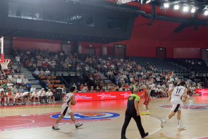 Eine Gruppe von Menschen, die Basketball in einer Turnhalle spielen, mit einem Basketballkorb auf der linken Seite und Zuschauern im Hintergrund.