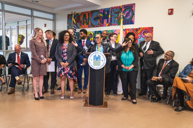 Gruppe von Menschen vor einem Podium mit Mikrofon und einer Tafel mit dem Text "Sancho-Stadtrat tritt dem Stadtrat bei", mit sitzenden und stehenden Gästen, Flaggen an der Rückwand und einem Feuerlöscher.