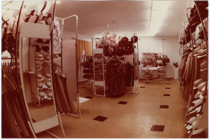 Altes Foto eines Kleiderladen-Interieurs mit Kleiderstangen, Hutständern, einem Spiegel, einem Stuhl, einem Tisch mit Gegenständen, Vorhängen und Deckenleuchten.