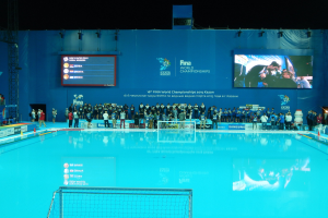 Gruppe von Menschen, die um ein Hallenbad mit Netz am Boden stehen, mit einer Wand im Hintergrund, die "FINA World Championships 2015 Kazan" auf einem Bildschirm anzeigt, begleitet von Trägern und Lichtern.